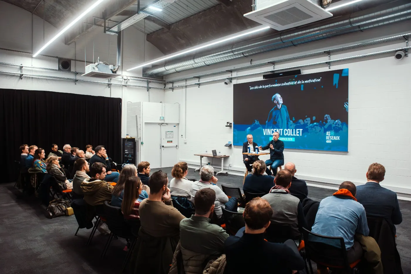 Salle comble lors d'une conférence, deux intervenants assis devant un écran de présentation